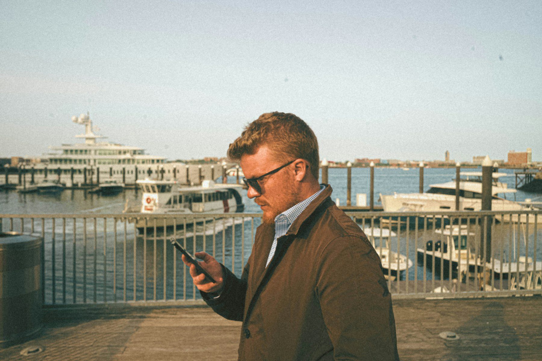 A man on a marina dock reading a message on his phone, boats moored behind him.